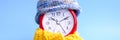 Red round alarm clock in knitted wool blue hat and yellow scarf on a blue background. winter time concept. winter season. cozy and Royalty Free Stock Photo
