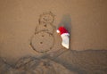 red Santa Claus hat on a white stone and a snowman painted on sand on coastal beach