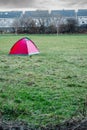 Red small tent on a green field and row of houses behind wall in the background. Homeless and successful people life concept.