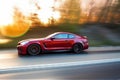 a red sports car driving down a road with trees in the backgrouund and the sun shining on the trees in the backgrouund