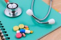 Red stethoscope, pencil and many colorful pills lying on a thin green book.