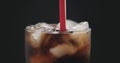 Red straw in cola with ice cubes in glass over black background