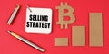On a red surface lie the bitcoin symbol, a pen and a notepad with the inscription - SELLING STRATEGY