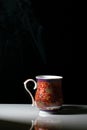 A red tea cup with designs on it placed on a white reflective table and smokes of hot tea coming out of it Royalty Free Stock Photo