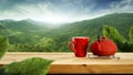 A red teapot on metal stand and a red mug on wooden table with beautiful green trees mountains and blue cloudy sky background. Royalty Free Stock Photo