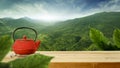 A red teapot on a metal stand on wooden table with beautiful green trees mountains and blue cloudy sky background. Royalty Free Stock Photo