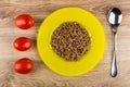 Red tomatoes, yellow plate with boiled buckwheat, spoon on table