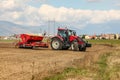 Red tractor sowing, pulling seeder trailer behind, on dry field, some houses and mountains in background Royalty Free Stock Photo