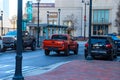 A red truck driving on a street lined with parked cars, tall black light posts and bare winter trees and buildings