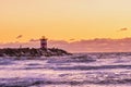 Red and white striped lighthouse during a vibrant sunset. Water hits the black boulders in waves. Dramatic sky in golden color. Royalty Free Stock Photo