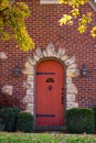 Red wood arched door surrounded by a rock frame in a brick house with castle like hardware with fall leaves hanging down on an aut