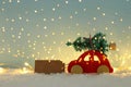 Red wooden car carrying a christmas tree over snow in front of blue background and golden garland lights. Royalty Free Stock Photo