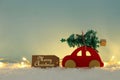 Red wooden car carrying a christmas tree over snow in front of blue background and golden garland lights. Royalty Free Stock Photo