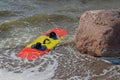Red and yellow kiteboarding board with carrying handle and slippers attached to it washed up by foaming wave next to large boulder