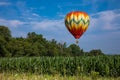 Red, Yellow, Orange, Green, and White Teardrop Shaped Hot Air Balloon Over Corn Field on Sunny Day with Trees and Cloudy Blue Sky Royalty Free Stock Photo