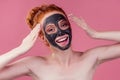 Redhaired ginger teenager girl with black clay mask on her pretty face on pink studio background