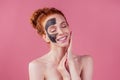 Redhaired ginger teenager girl with black clay mask on her pretty face on pink studio background