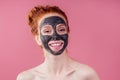 Redhaired ginger teenager girl with black clay mask on her pretty face on pink studio background