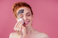 Redhaired ginger teenager girl remove with cotton black clay mask on her pretty face on pink studio background