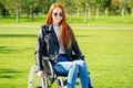 Redhaired ginger woman feeing happy,she sitting wheelchair,wearing sunglasses and warm leather jacket in summer park