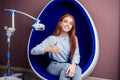 Redhaired ginger young cheerful female client sitting in dental office and getting smile whitening in egg chair Royalty Free Stock Photo