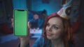 A redhaired girl in a festive hat shows a smartphone with a green screen, smiles. Close up of a girl with a smartphone Royalty Free Stock Photo