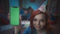 A redhaired girl in a festive hat shows a smartphone with a green screen, smiles. Close up of a girl with a smartphone Royalty Free Stock Photo