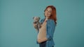 Redhaired young pregnant woman holding a teddy bear. Woman with a toy in her hands in the studio on a blue background