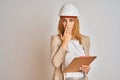 Redhead caucasian business woman wearing safety helmet and holding clipboard cover mouth with hand shocked with shame for mistake,