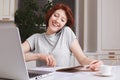 Redhead cheerful woman being busy with work, recieves orders on smart phone, makes notes in spiral notebook, glad to have many vis Royalty Free Stock Photo