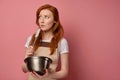 A redhead girl in a apron and a T-shirt stands on a pink background, with a bowl and a whisk in hands and looks scared Royalty Free Stock Photo