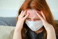 A redhead girl in a medical mask has a headache. Woman with a sore head sits on the bed Royalty Free Stock Photo