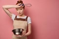 The redhead girl, soiled in flour, in an apron, stands on a pink background and looks puzzled at the bowl, with hand to