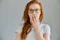 Redhead girl in a white T-shirt and glasses stands on a gray background, looks surprised into the frame, covering mouth Royalty Free Stock Photo