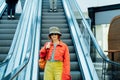 Redhead hipster young woman in bright clothes, sun glasses and bucket hat with backpack bag standing on escalator going