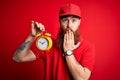 Redhead Irish delivery man with beard holding alarm clock for deadline over red background cover mouth with hand shocked with Royalty Free Stock Photo