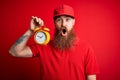 Redhead Irish delivery man with beard holding alarm clock for deadline over red background scared in shock with a surprise face,