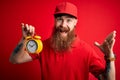 Redhead Irish delivery man with beard holding alarm clock for deadline over red background very happy and excited, winner Royalty Free Stock Photo