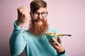 Redhead Irish healthy man with beard eating vegetarian green salad over pink background annoyed and frustrated shouting with