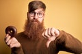Redhead Irish man with beard eating chocolate donut over yellow background with angry face, negative sign showing dislike with