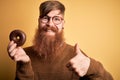 Redhead Irish man with beard eating chocolate donut over yellow background happy with big smile doing ok sign, thumb up with Royalty Free Stock Photo