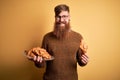 Redhead Irish man with beard eating french croissant pastry over yellow background with a happy face standing and smiling with a