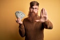 Redhead Irish man with beard holding a bunch of 1 dolla banknotes over yellow background with open hand doing stop sign with