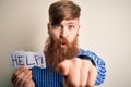 Redhead Irish man with beard holding paper with help message over blue background pointing with finger to the camera and to you,