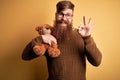 Redhead Irish man with beard hugging teddy bear stuffed animal over yellow isolated background doing ok sign with fingers,