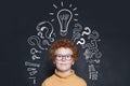 Redhead kid with lightbulb and question marks on blackboard background. Brainstorming and idea concept