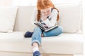 Redhead little girl sitting on sofa and reading book Royalty Free Stock Photo