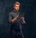 Redhead man dressed in an elegant vest with tie and kilt using a tablet in studio against a dark textured wall