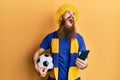 Redhead man with long beard football hooligan cheering game holding smartphone angry and mad screaming frustrated and furious,