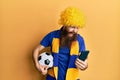 Redhead man with long beard football hooligan cheering game holding smartphone angry and mad screaming frustrated and furious,